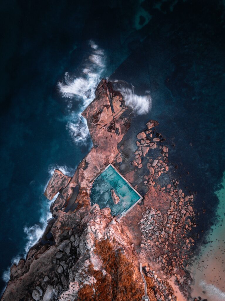 Aerial view of ocean waves crashing on rocks around a natural coastal pool with textured terrain Wallpaper