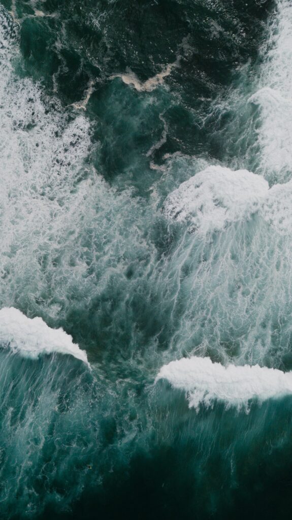 Aerial view of dynamic ocean waves with swirling white foam over deep blue-green water background Wallpaper