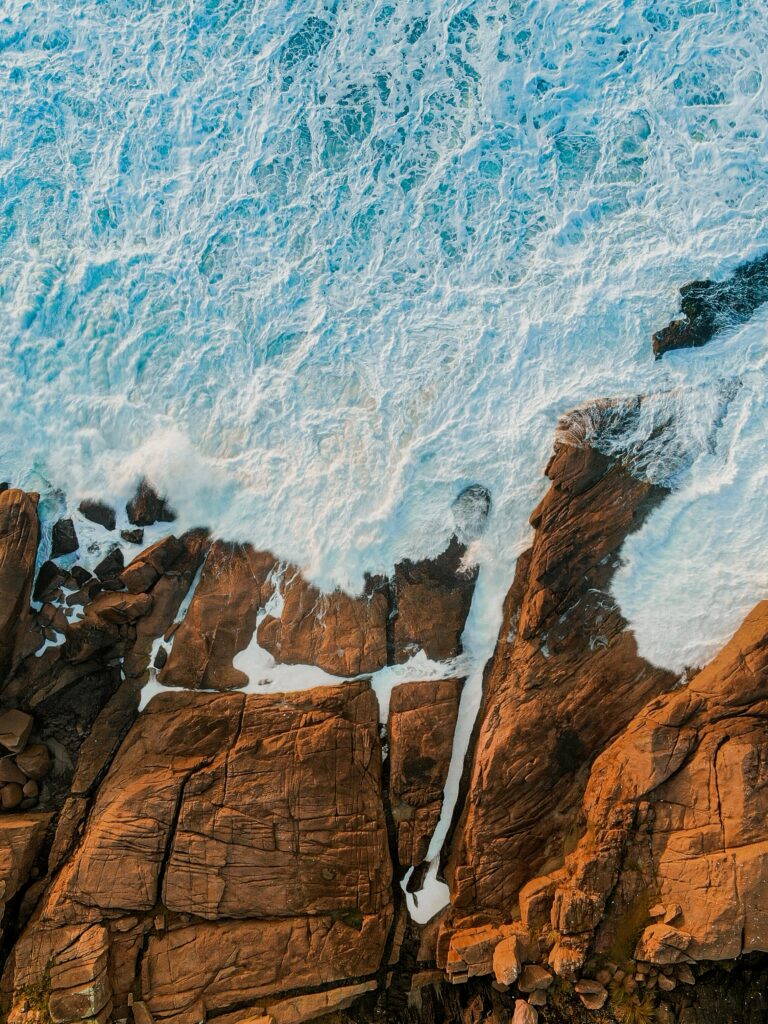 Aerial view of rugged brown rocky coast with crashing foamy blue waves and textured shoreline Wallpaper