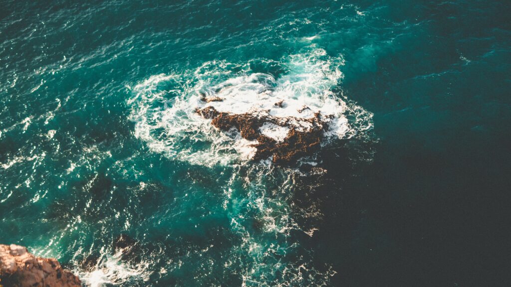 Aerial view of ocean waves crashing rugged rocks with white spray against turquoise water background Wallpaper