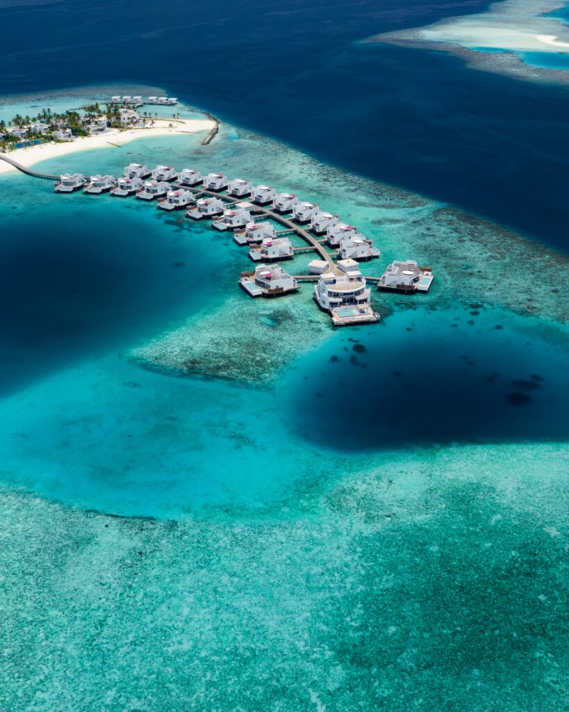 Aerial view of curved overwater bungalows above turquoise ocean near pristine island and blue sky Wallpaper