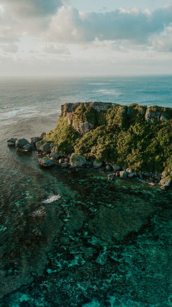 Aerial view of rocky island with lush greenery surrounded by turquoise sea and coral reefs Wallpaper