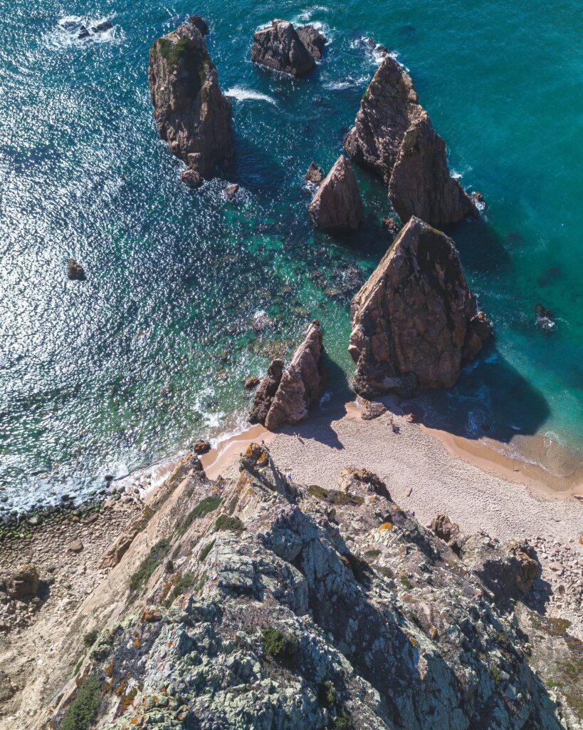 Aerial view of rugged coastal rocks and turquoise sea with waves on sandy shore under sunlight Wallpaper