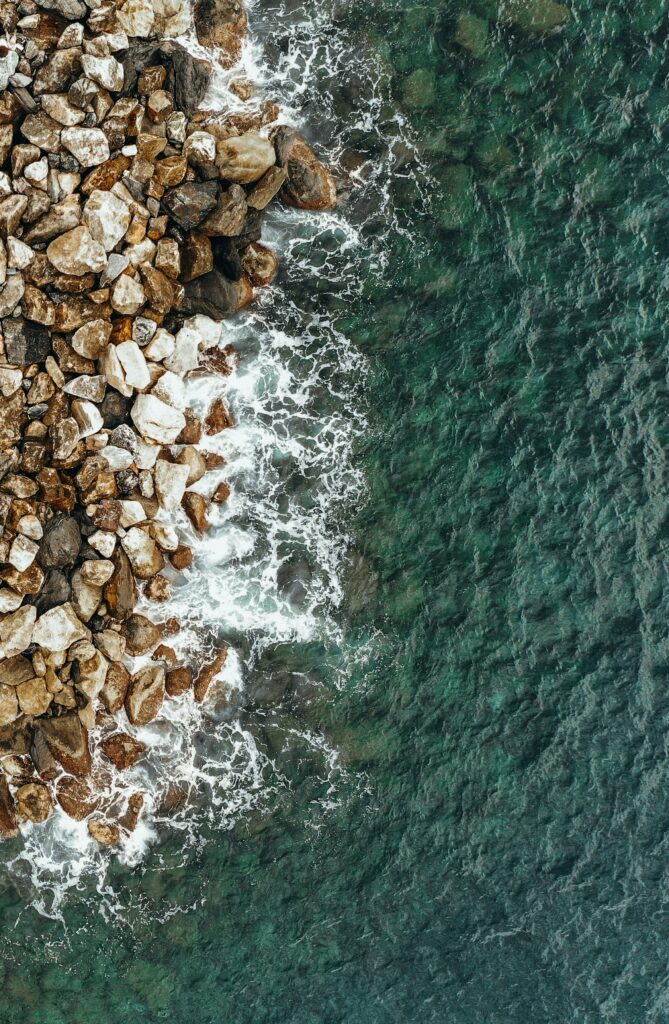 Aerial view of rugged coastal stones meeting vibrant blue sea with foamy crashing waves Wallpaper