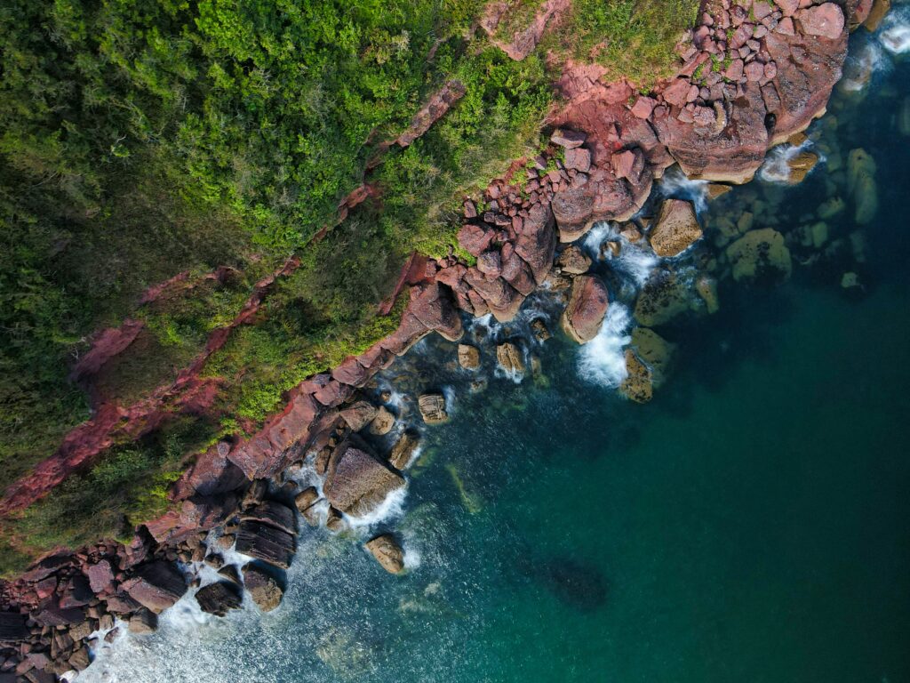 Aerial view of rugged coastline with green vegetation red rocks and blue green waves crashing shore Wallpaper