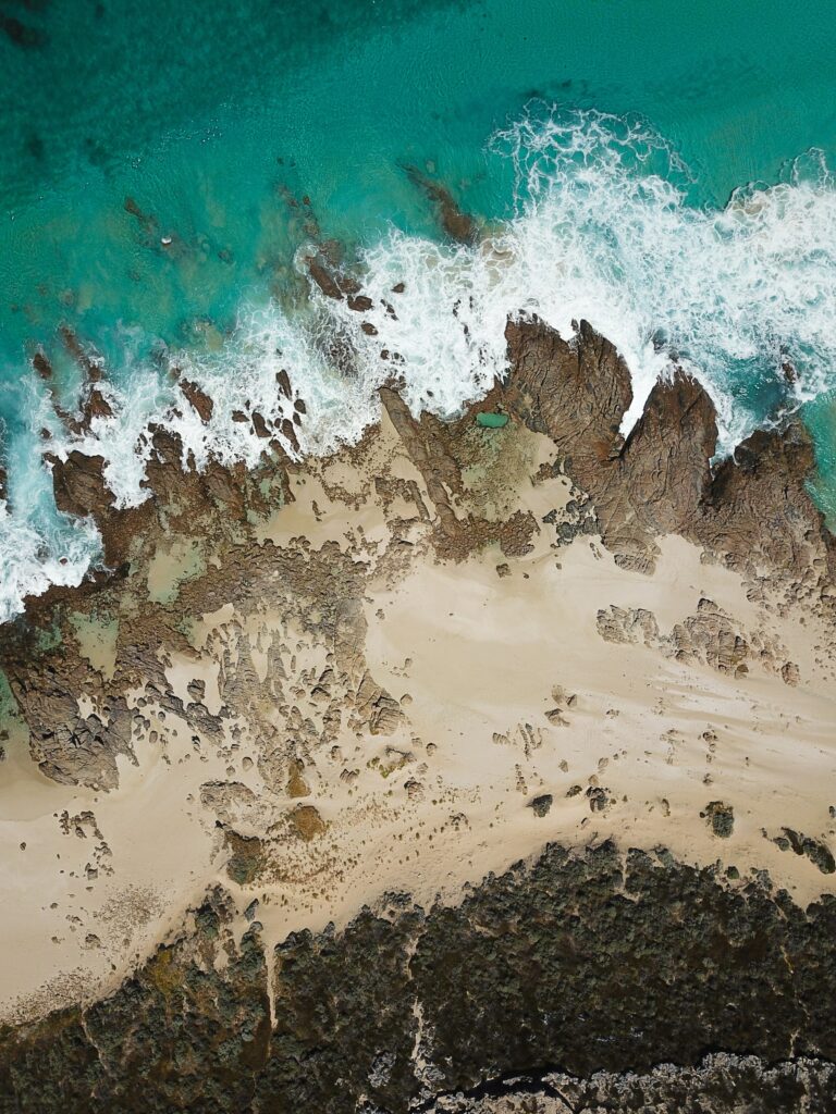 Aerial view of rugged coastline with turquoise waves crashing on rocky outcrops and sandy beach Wallpaper