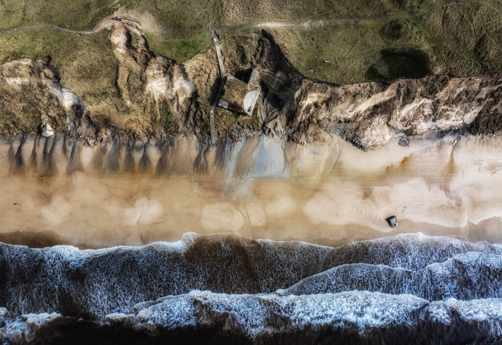 Aerial view of rugged coastline with waves, sandy beach, cliffs, and green grassy terrain wallpaper