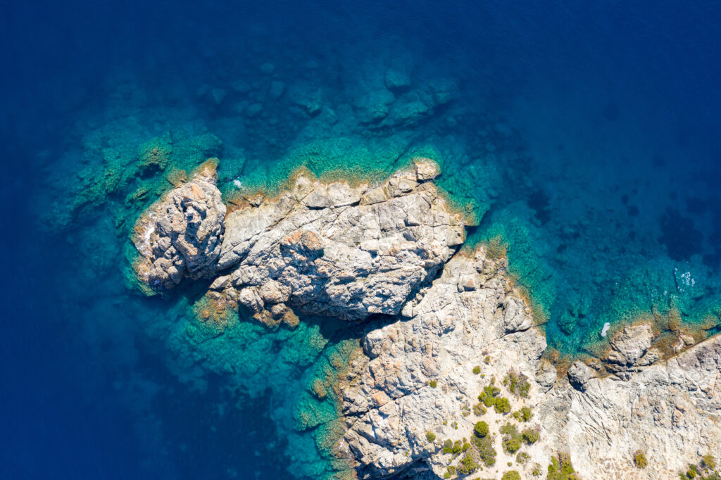 Aerial view of rugged rocks and small island surrounded by vibrant turquoise to navy blue ocean water Wallpaper
