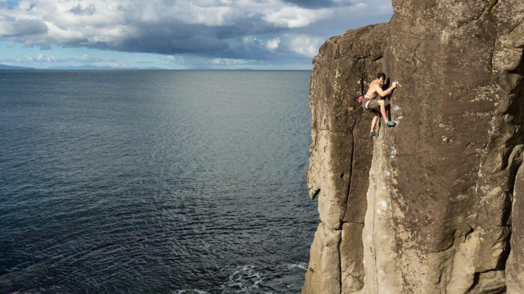 Aerial view of topless man rock climbing steep cliff above ocean showcasing strength and coast Wallpaper