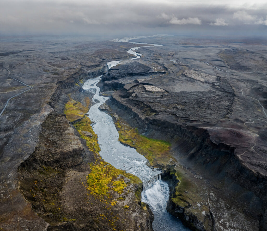 Aerial view of winding river through rocky terrain with green vegetation under dramatic clouds Wallpaper