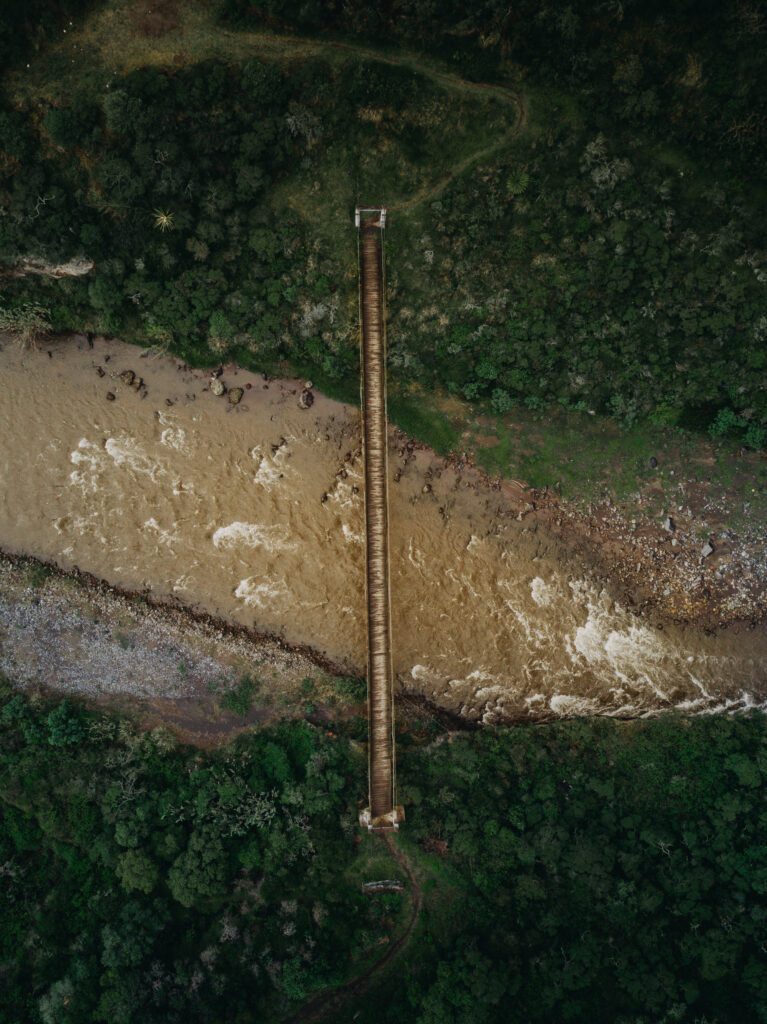 Aerial view of narrow bridge over muddy river surrounded by dense green forests wallpaper background