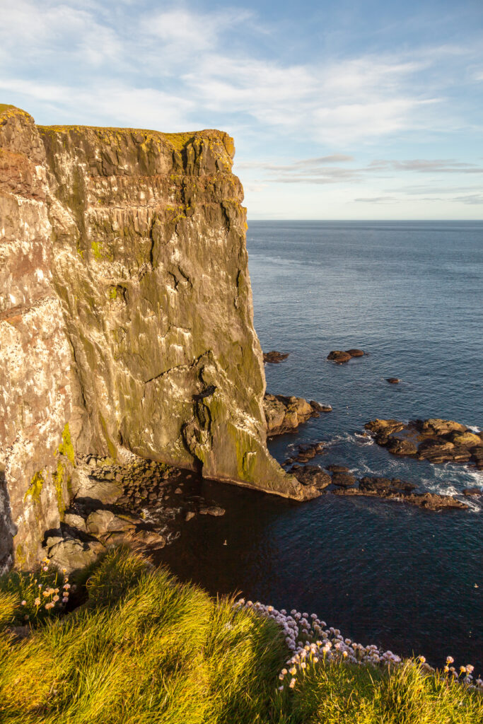 Aerial view of rugged rock cliff with green grass overlooking calm blue sea under sunlight Wallpaper