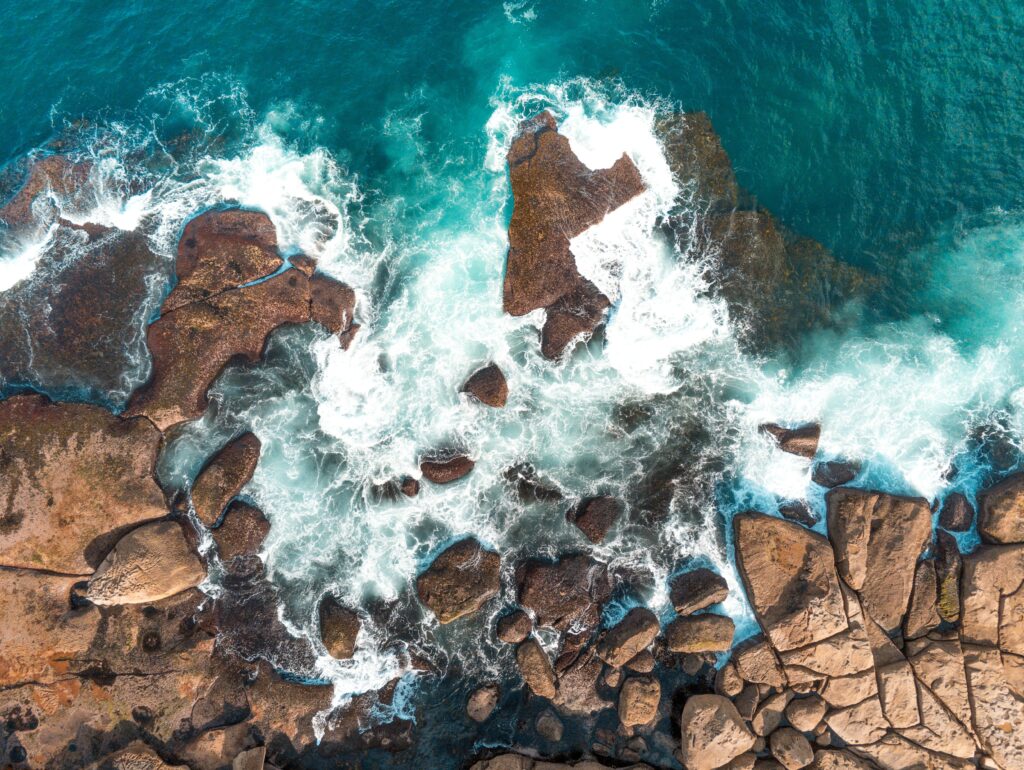 Aerial view of rugged coastal rocks with turquoise sea and white waves crashing on shore wallpaper