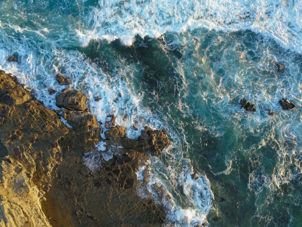 Aerial view of rugged rocky shoreline with crashing ocean waves white foam and turquoise water Wallpaper
