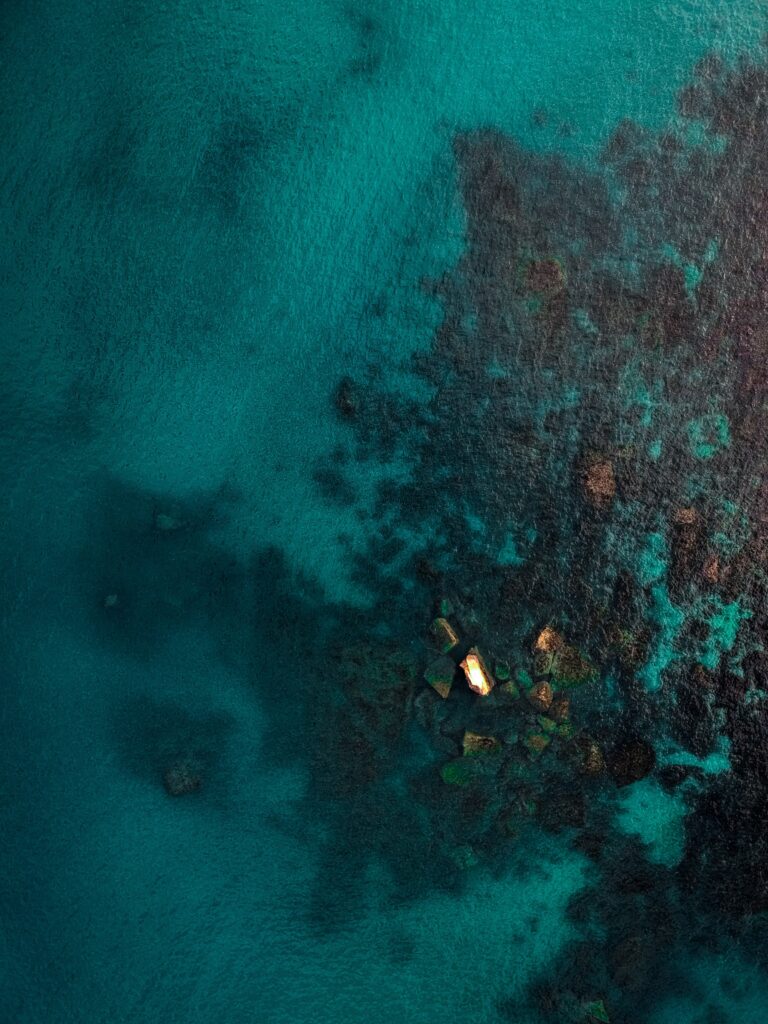 Aerial view of teal turquoise ocean with submerged stones and sunlight highlighting rocks Wallpaper
