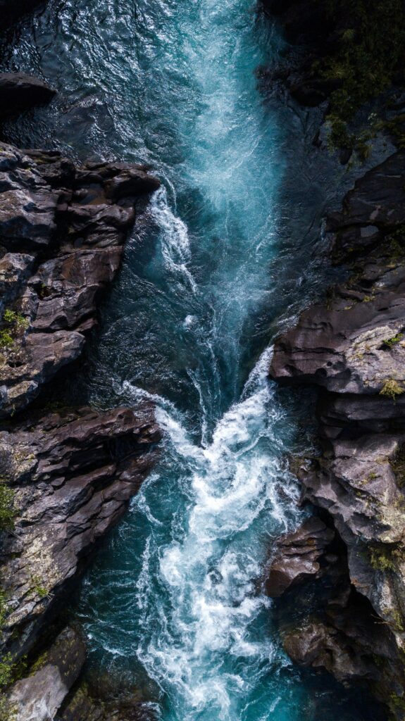 Aerial view of foamy turquoise water flowing through rugged rocks and dark stones wallpaper