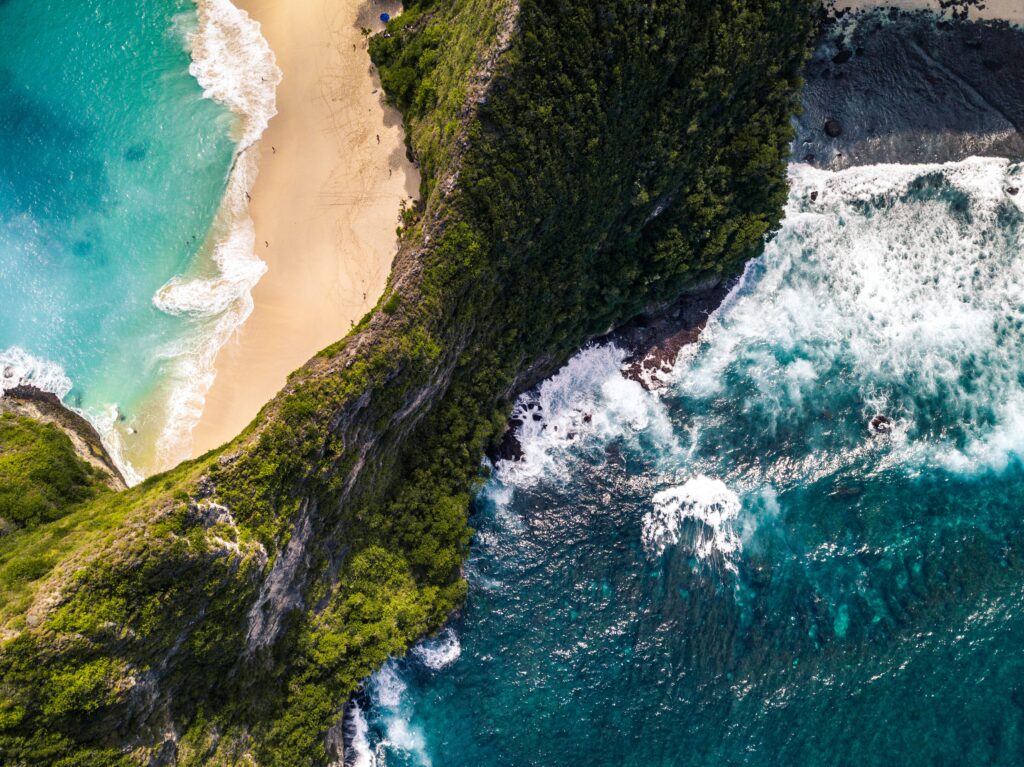 Aerial view of turquoise waves meeting golden sand with rocks and lush green coastal scenery Wallpaper