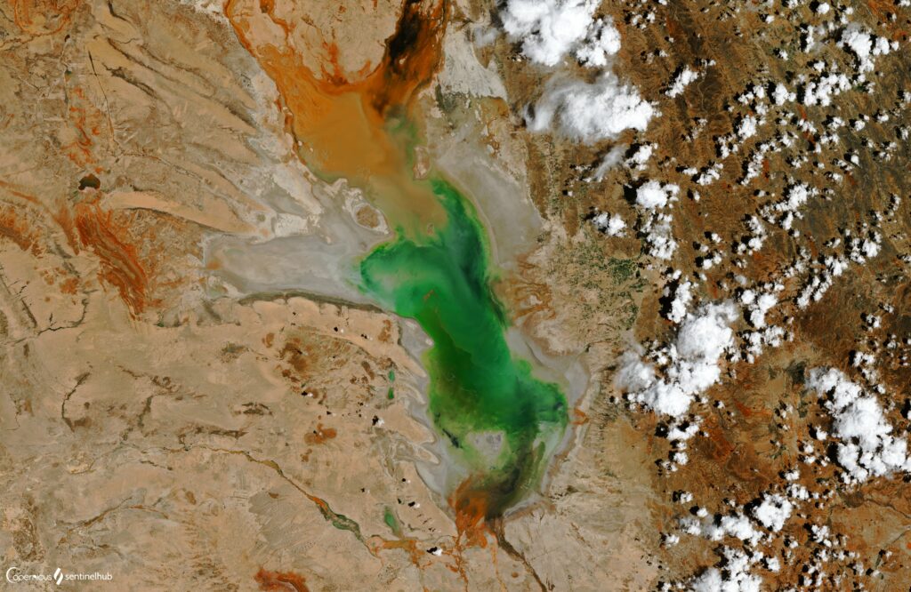 Aerial view of vibrant green turquoise lake surrounded by rugged brown orange terrain and clouds Wallpaper