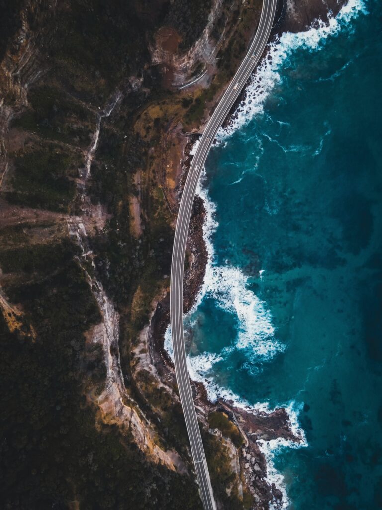 Aerial view of winding coastal road beside rugged cliffs lush greenery and vibrant blue sea waves Wallpaper