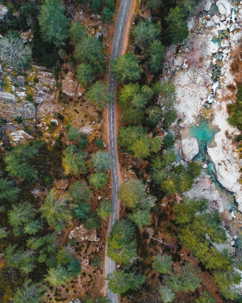 Aerial view of winding road through lush green forest beside turquoise stream and rocky terrain Wallpaper