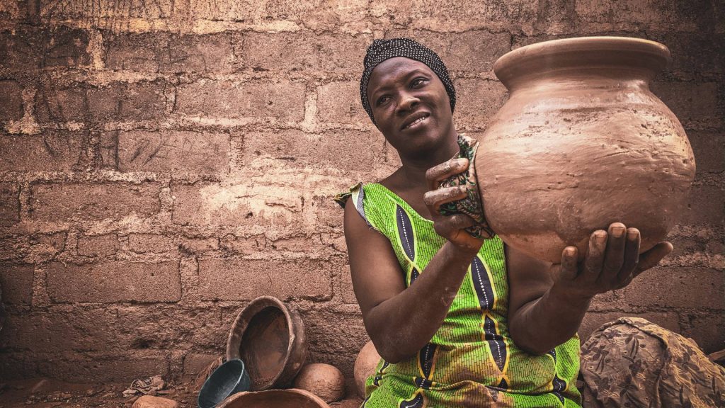 African woman in green dress holding handmade clay pot against rustic block wall wallpaper background