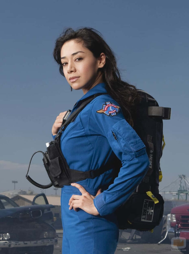 Aimee Garcia in blue rescue uniform smiling outdoors with backpack against wallpaper background