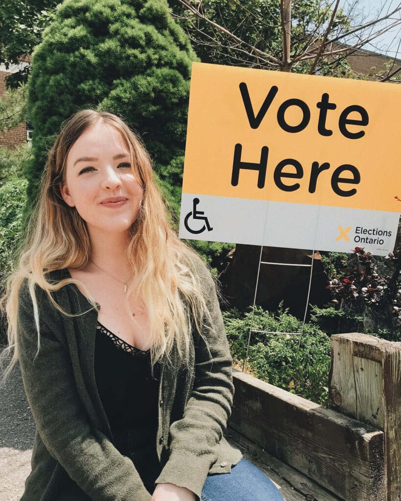 Aislinn Paul smiling warmly by Vote Here sign encouraging voting positive wallpaper background