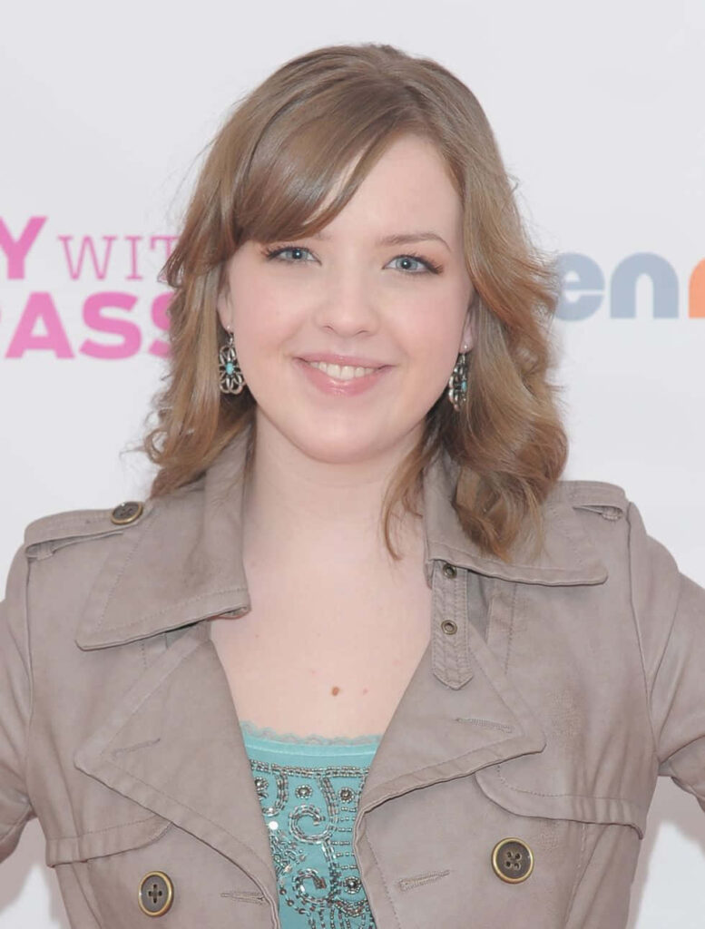 Aislinn Paul smiling confidently in beige jacket and blue top at public event wallpaper background