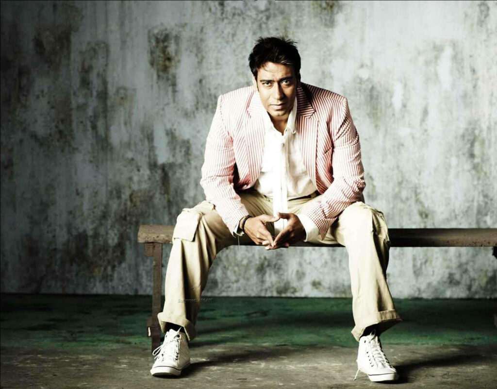 Bollywood actor Ajay Devgn stylishly seated in striped jacket and white shirt on textured backdrop Wallpaper