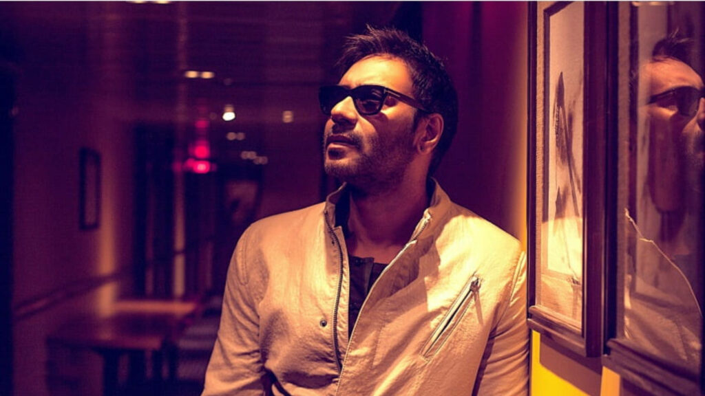 Ajay Devgn in light jacket and sunglasses standing confidently in warmly lit hallway with wallpaper