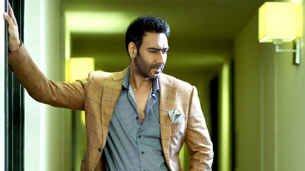 Ajay Devgn stylish in orange checkered suit and patterned shirt standing confidently hallway wall Wallpaper
