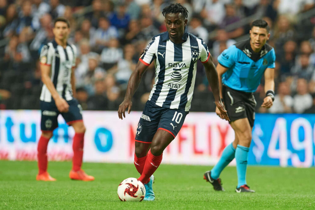 Aké Arnaud Loba dribbling soccer ball in intense match with players and referee background Wallpaper