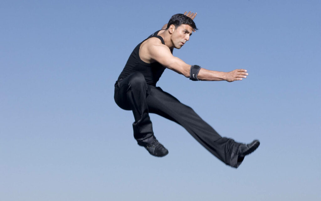 Akshay Kumar performing dynamic aerial kick against clear blue sky action wallpaper background