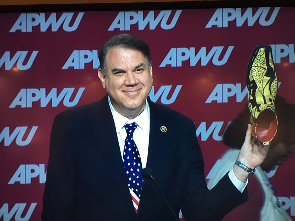 Alan Grayson American politician smiling holding decorative shoe at APWU event with red backdrop Wallpaper