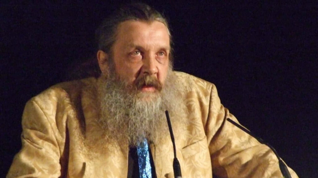 Alan Moore legendary author with long hair and beard posing against dark wallpaper background