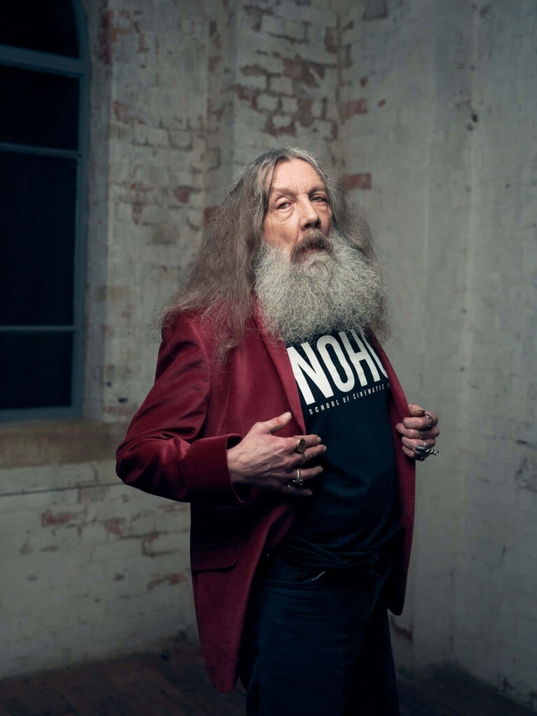 Portrait of British writer Alan Moore with long hair red blazer and graphic t-shirt against brick wall Wallpaper