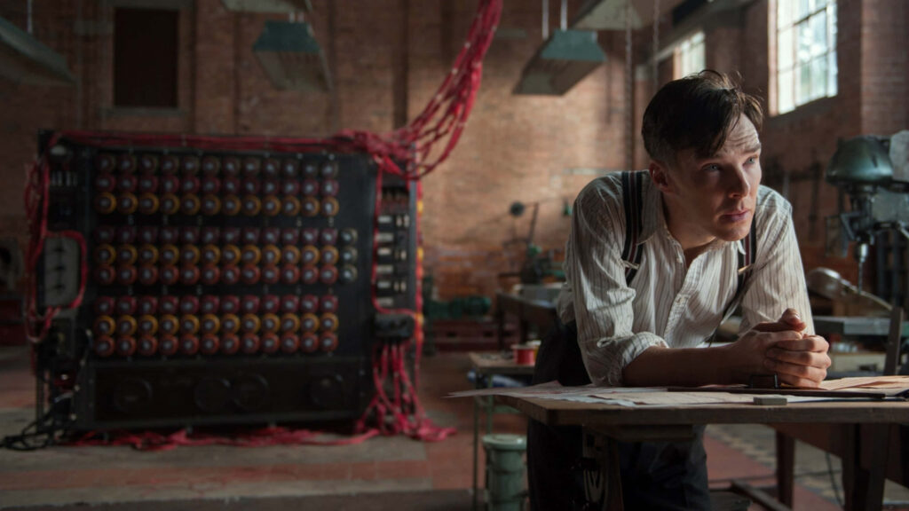 Alan Turing leaning over desk with Enigma machine in dim room iconic scene from The Imitation Game Wallpaper