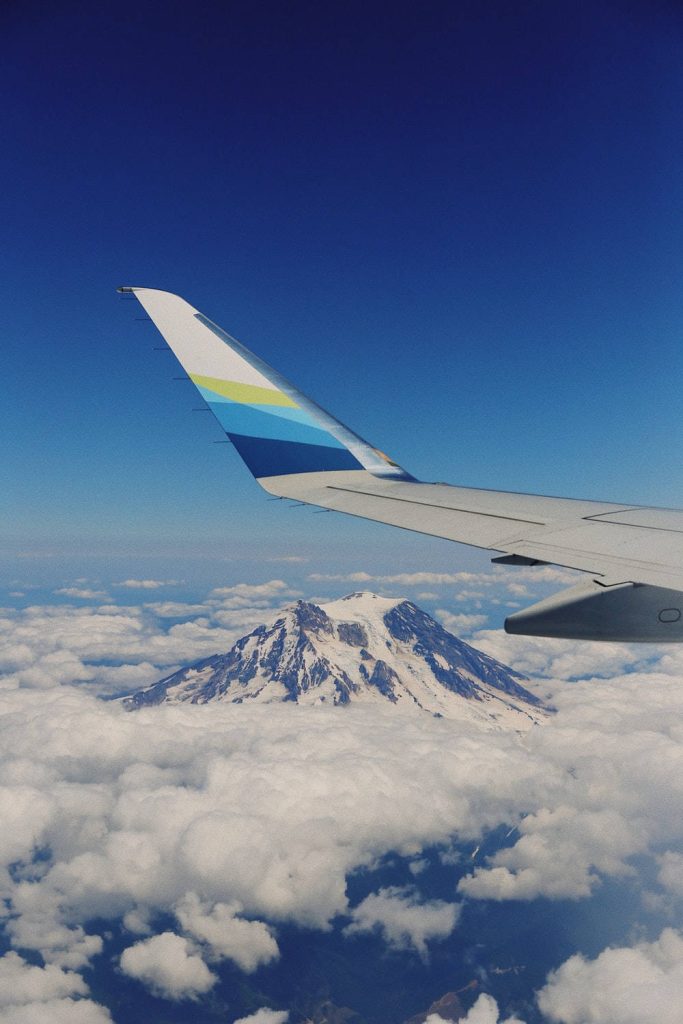 Alaska Airlines winglet flying over snowy Mount Rainier through clouds under blue sky wallpaper