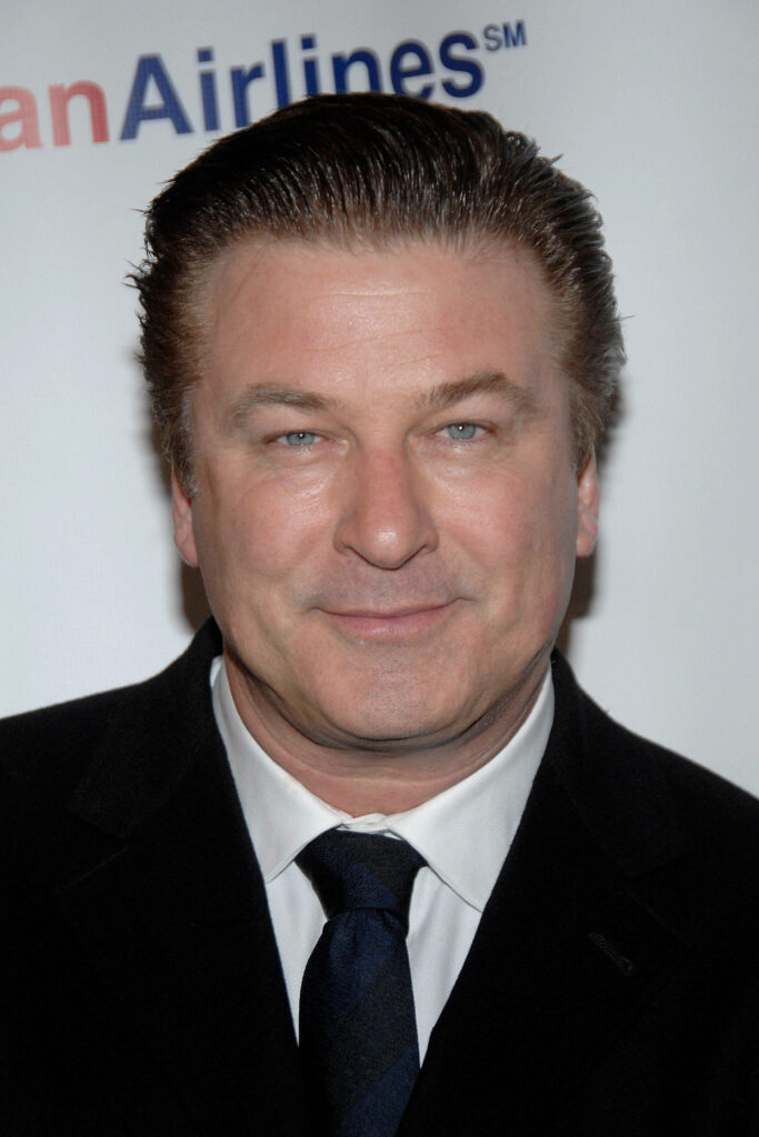 Alec Baldwin confident in dark suit and tie with smooth skin against soft wallpaper background