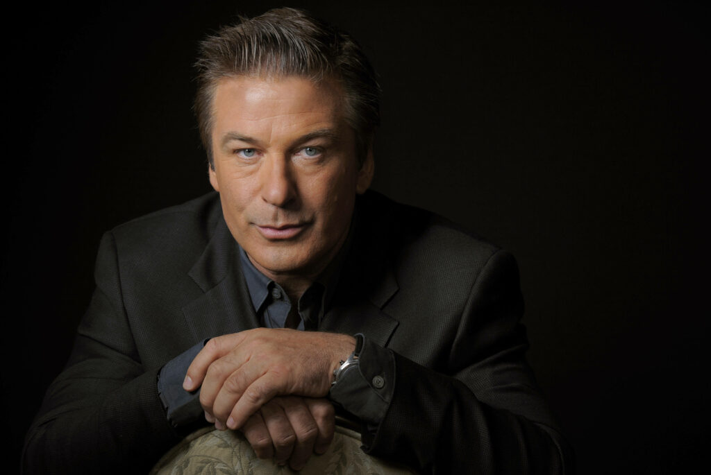 Alec Baldwin in dark suit confidently gazing serious against black wallpaper background