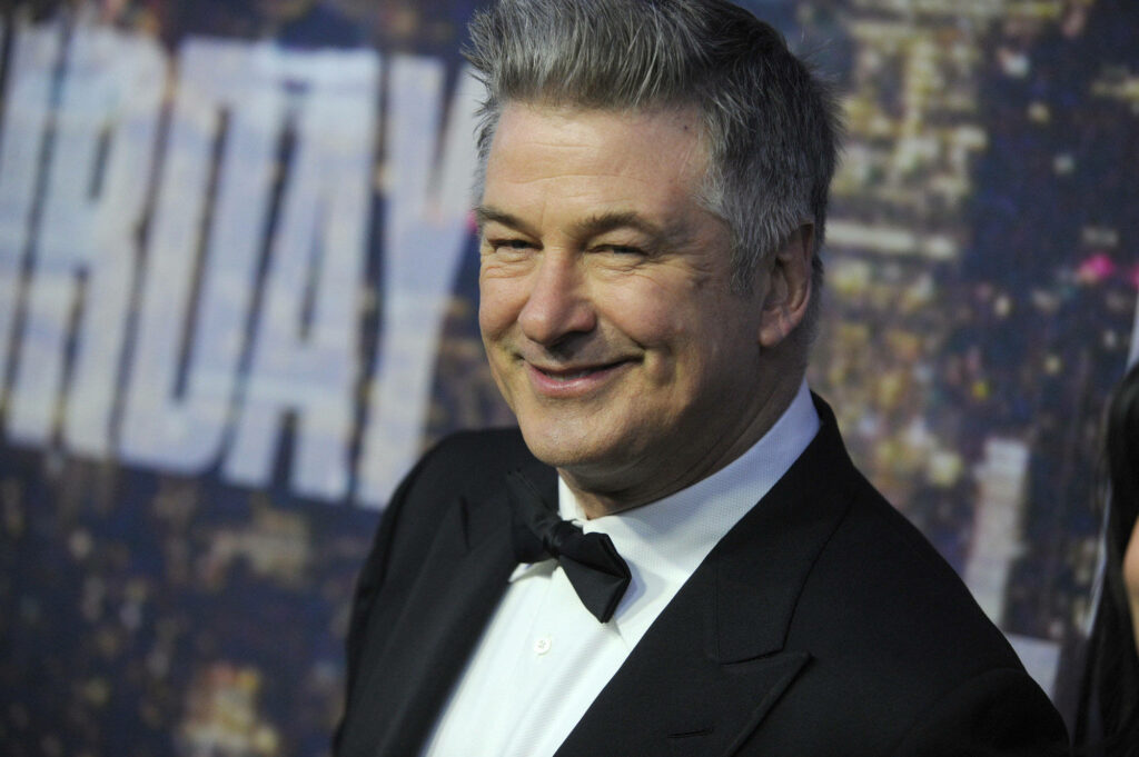Alec Baldwin in sharp black tuxedo smiling confidently with vibrant city skyline background Wallpaper