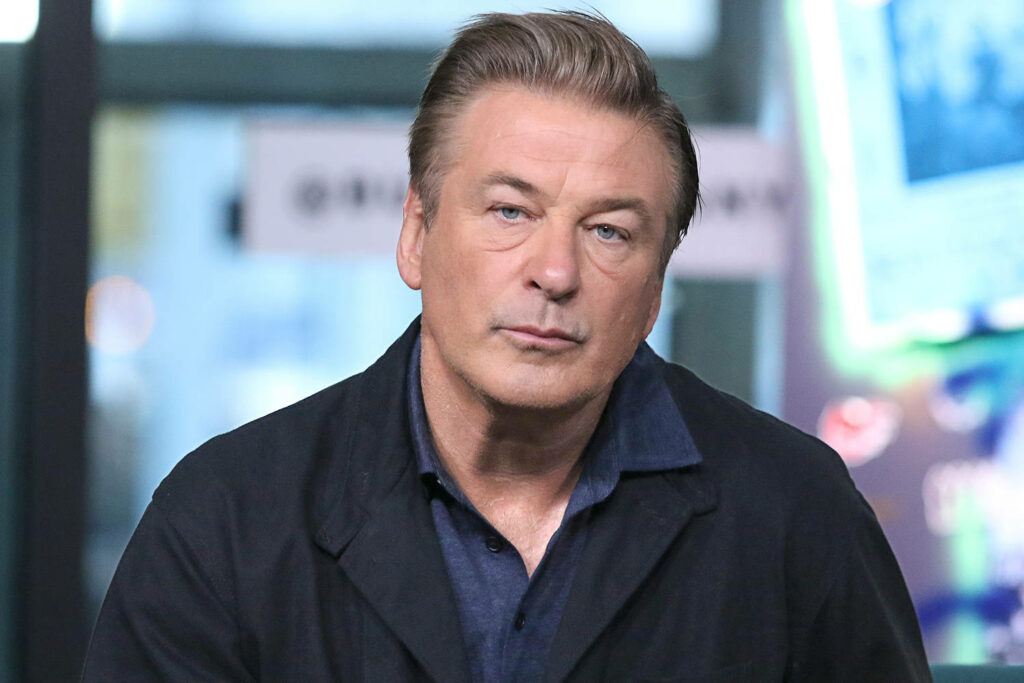 Alec Baldwin in dark jacket looking thoughtful during interview with blurred wallpaper background