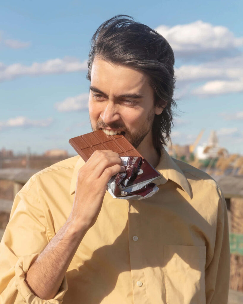 Alex G enjoying chocolate bar outdoors under bright sky relaxed candid wallpaper background