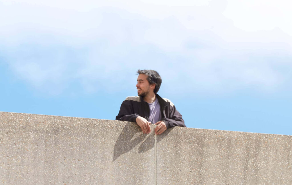 Alex G leaning casually on textured wall under clear blue sky calm relaxed wallpaper background