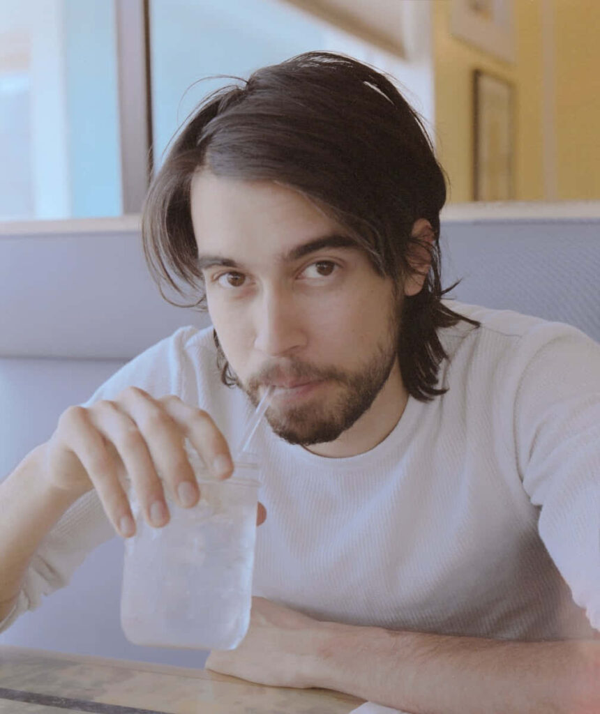 Alex G relaxed at cafe table sipping water from glass jar soft cozy atmosphere wallpaper background