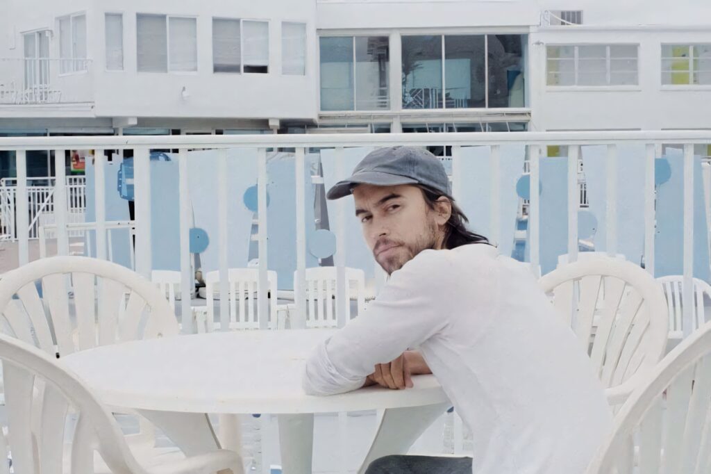 Alex G seated at seaside balcony white table thoughtful gaze pastel tones modern buildings background Wallpaper