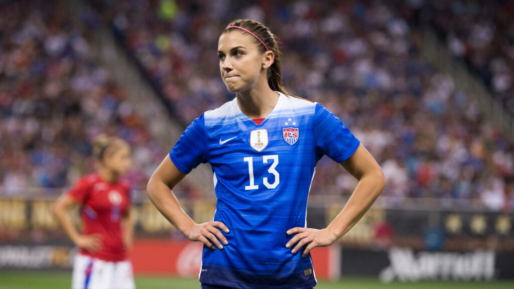 Alex Morgan focused in USA soccer jersey during intense game in packed vibrant stadium background Wallpaper
