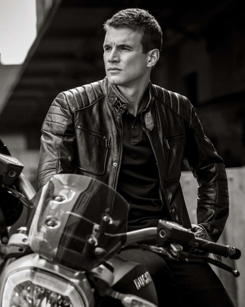 Alex Russell in sleek leather jacket sitting on motorcycle monochrome wallpaper background