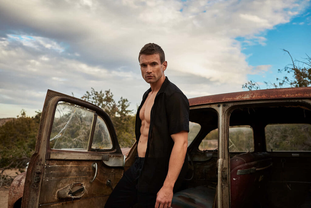 Alex Russell stands beside weathered vintage car in desert landscape rugged adventurous wallpaper