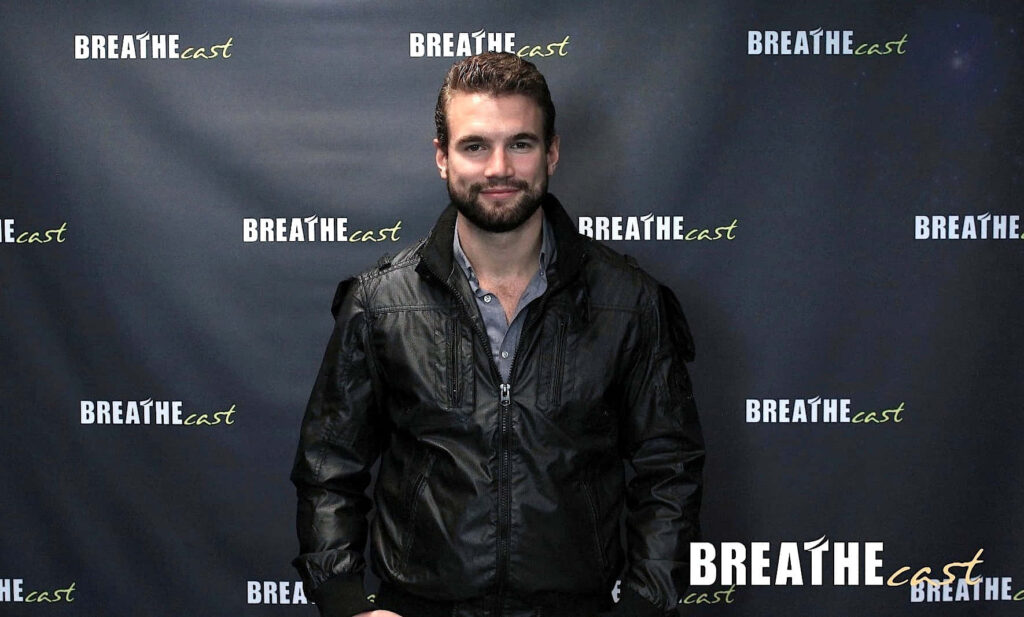 Alex Russell poses confidently at Breathe Cast event in stylish black jacket against wallpaper backdrop